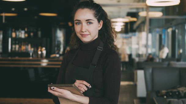 Portrait of cheerful and beautiful young waitress standing, keeping menu, and smiling sincerely in slylish cafe. Bar counter with many hard and soft drinks is in the background - Zdjęcie, obraz