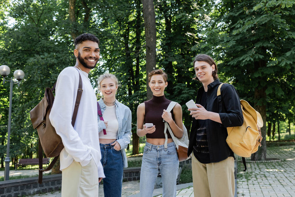 Smiling interracial students with backpacks and smartphones looking at camera in park  - 写真・画像