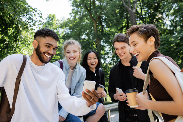 Estudiante afroamericano sosteniendo smartphone cerca de amigos interracial en el parque  - Foto, imagen