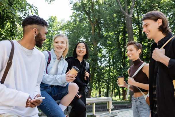 Positive african american student using smartphone near interracial friends with coffee in park  - 写真・画像