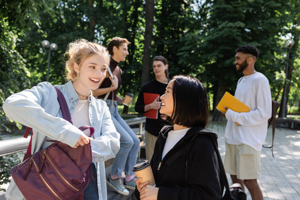 Positive interracial students with coffee to go talking near blurred friends in park  - Photo, Image