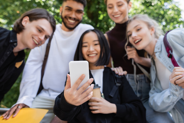 Blurred asian student holding smartphone near interracial friends in park  - Foto, Bild