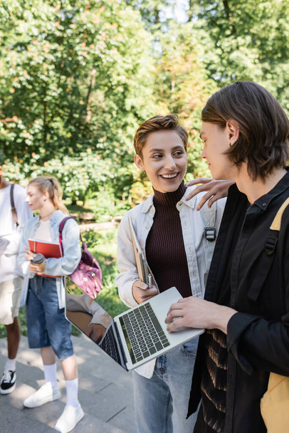 Positive students with laptop talking near blurred multiethnic friends in park  - Foto, Bild