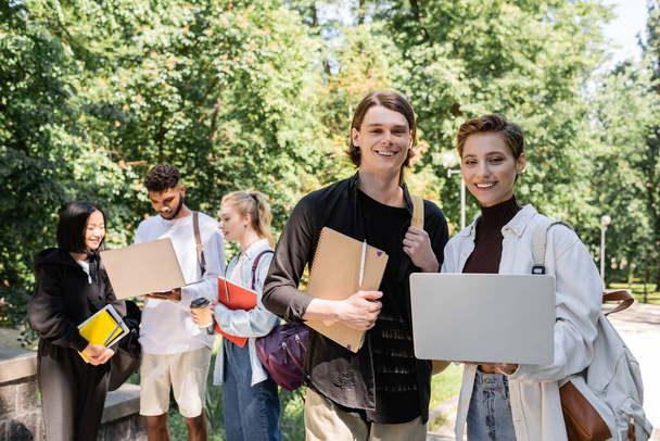 Smiling students with laptop looking at camera near blurred multiethnic friends in park  - Photo, image