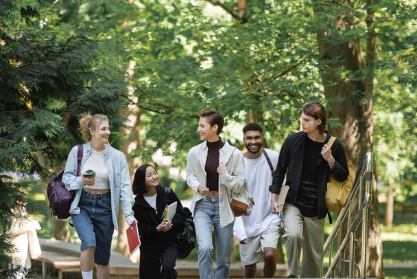Cheerful students with backpacks walking near multiethnic friends in park  - Fotó, kép