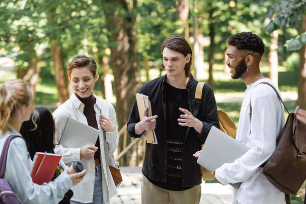 Positive interracial students with laptops talking to blurred friends in park  - Photo, image