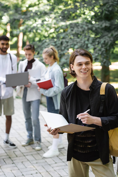 Smiling student holding notebook near blurred interracial friends in park  - Foto, Imagem