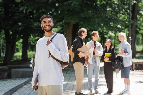 Smiling african american student with laptop standing near blurred friends in summer park  - Photo, Image