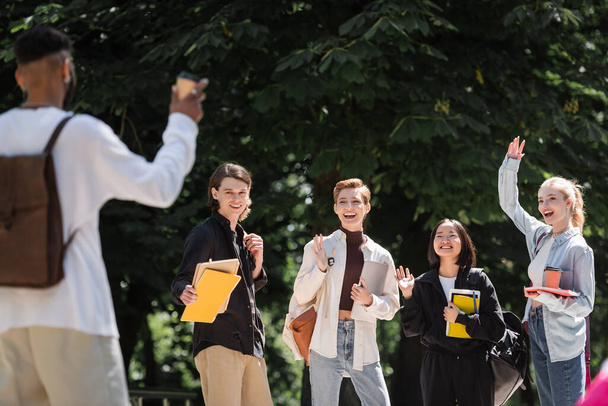 Позитивні міжрасові студенти махають руками поблизу розмитого афроамериканського друга в парку
  - Фото, зображення