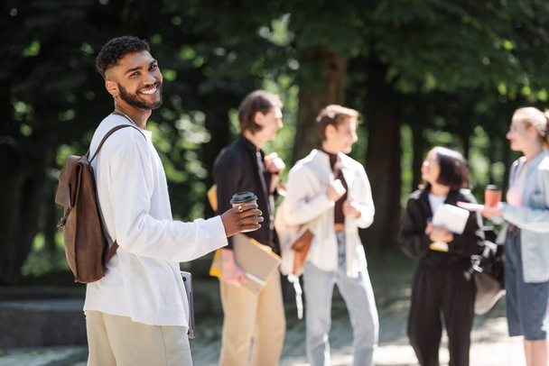 Smiling african american student holding coffee to go near blurred friends in park  - Foto, imagen