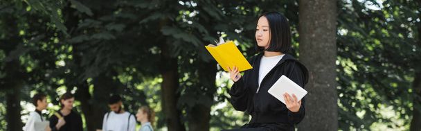 Азійський студент тримає в парку цифрову табличку і книжку для читання.  - Фото, зображення