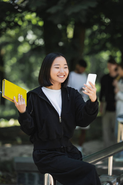 Sonriendo asiática estudiante celebración de libro y tomando selfie en smartphone en parque - Foto, Imagen