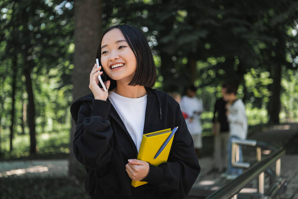 Positive asian student holding book and talking on smartphone in park  - Foto, Imagem