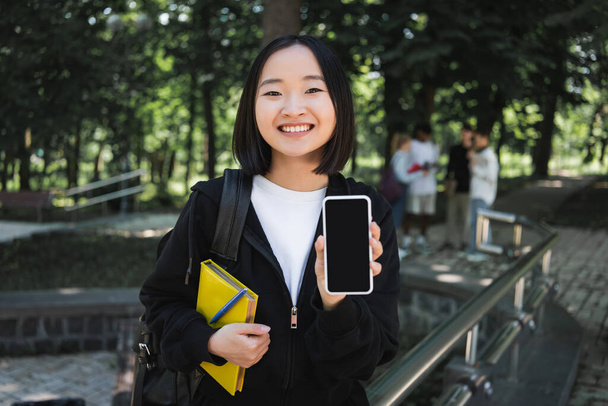 Весёлый азиатский студент с книгой, показывающей смартфон в парке  - Фото, изображение