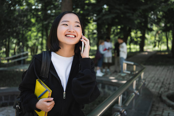 Happy asian student with book and backpack talking on smartphone in park  - 写真・画像