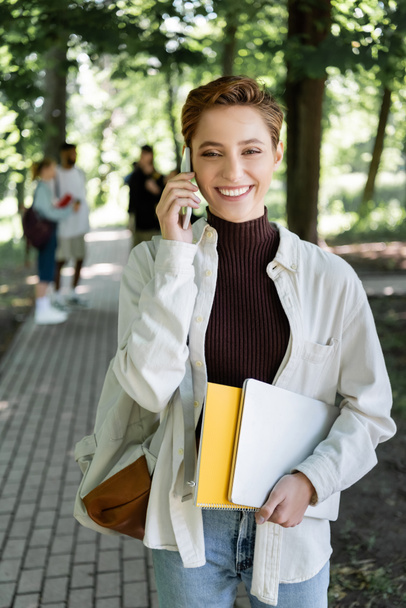 Positive student with laptop and notebook talking on smartphone in park  - Photo, image