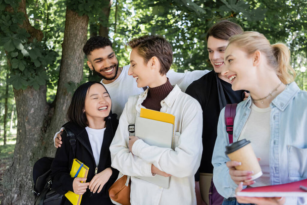 Cheerful multicultural students with copy books talking in park  - 写真・画像