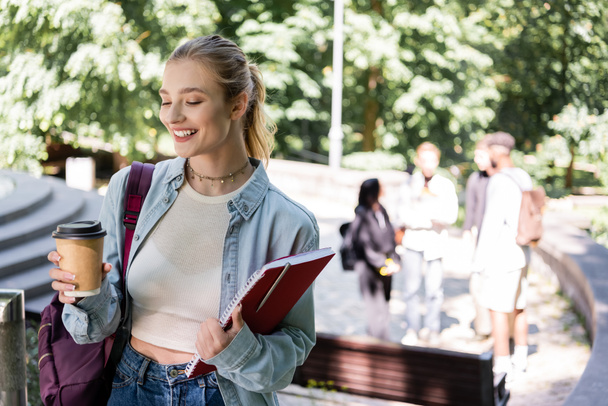Smiling student holding coffee to go and notebook in park  - Фото, зображення