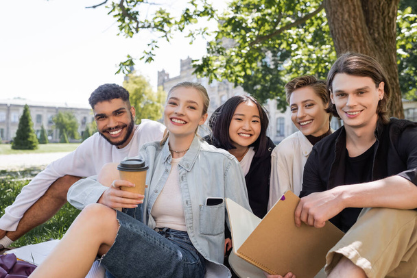 Позитивные мультикультурные студенты с ноутбуком и кофе, смотрящие в камеру на траве в парке  - Фото, изображение