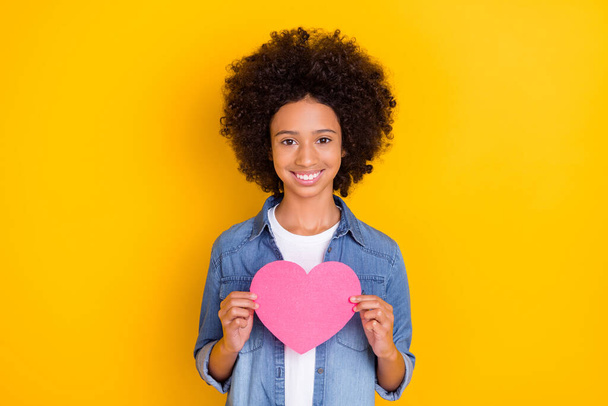 Portrait of beautiful trendy cheery teen-aged girl holding in hands pink heart isolated over bright yellow color background. - Φωτογραφία, εικόνα