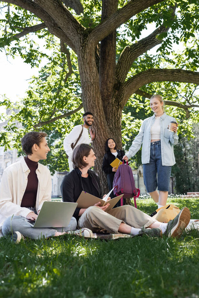 Estudante alegre com bebida takeaway em pé perto de amigos multiétnicos na grama no parque de verão  - Foto, Imagem