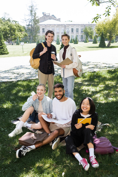 Cheerful multiethnic students with notebooks and coffee looking at camera on grass in park  - Fotó, kép