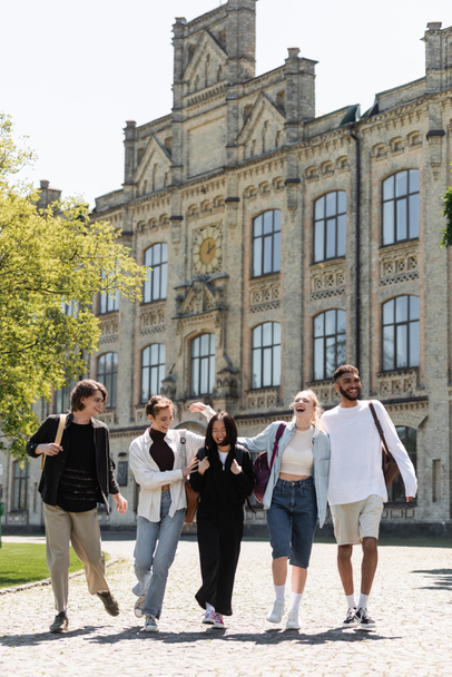 Позитивні багатоетнічні студенти з рюкзаками, що ходять поблизу розмитого університету на відкритому повітрі
  - Фото, зображення