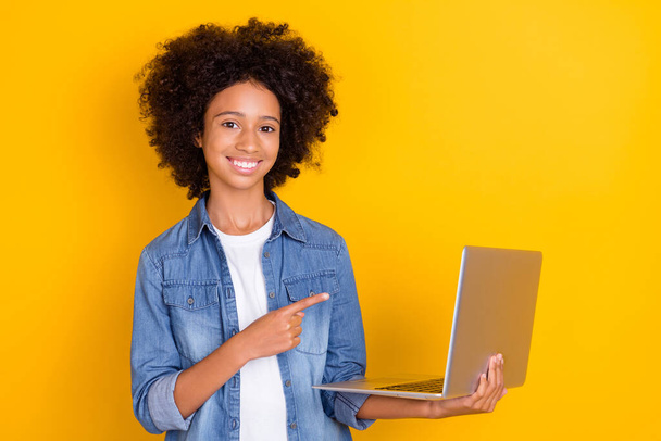 Portrait of beautiful trendy cheerful teen girl holding demonstrating laptop isolated over bright yellow color background. - Foto, Imagem
