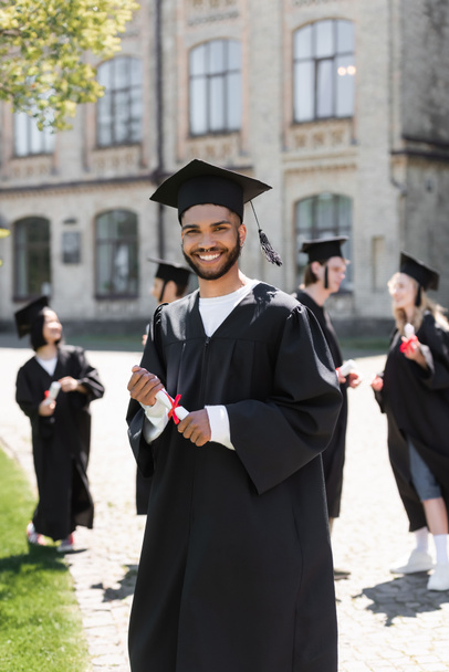 Young african american bachelor in gown holding diploma and looking at camera in park  - Valokuva, kuva