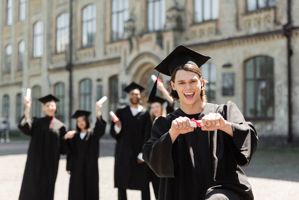 Positive bachelor holding diploma and looking at camera near blurred interracial friends outdoors  - Foto, Imagem