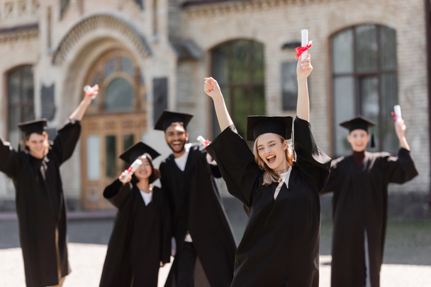 Excited student in bachelor gown holding diploma near multiethnic friends outdoors  - Fotoğraf, Görsel