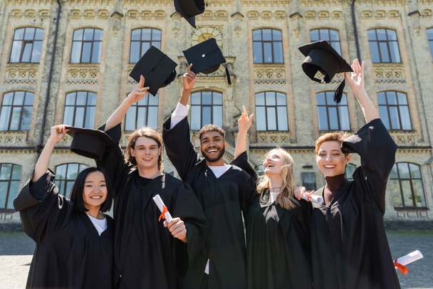 Smiling multiethnic bachelors holding diplomas and throwing caps outdoors  - Valokuva, kuva