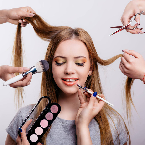 Beautiful girl surrounded by hands of makeup artists and hair stylist with brushes and scissors near her face. Young woman in a beauty salon. - Valokuva, kuva