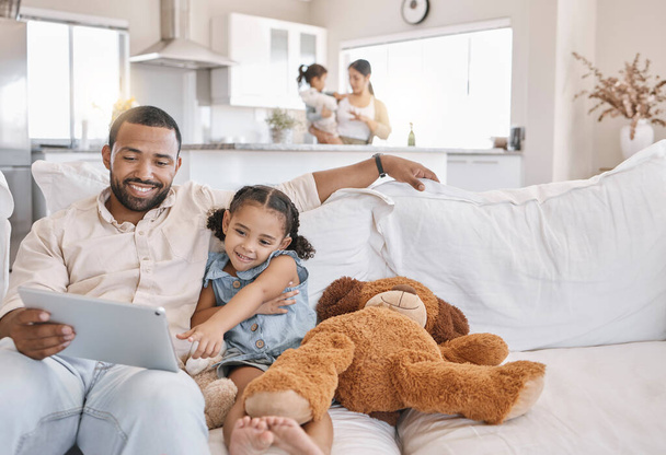 Gemengd ras vader en dochter ontspannen op de bank in de woonkamer met behulp van een digitale tablet, terwijl zijn vrouw zorgt voor hun andere dochter in de keuken. - Foto, afbeelding