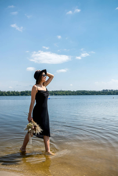 Pretty woman walking along barefoot in evening black dress and black hat  in city lake. lifestyle - Фото, зображення