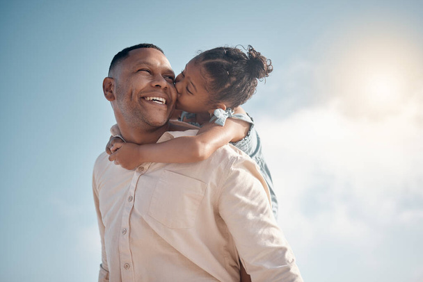 Smiling mixed race single father carrying little daughter on piggyback with copyspace. Adorable, happy, hispanic girl bonding with parent and kissing cheek on beach. Man and child enjoying free time. - Foto, imagen