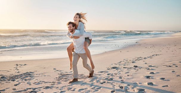 Happy couple playing on the beach. Carefree young couple having fun on holiday together. Husband giving his wife a piggyback ride. Married couple being playful on vacation together. - Foto, immagini
