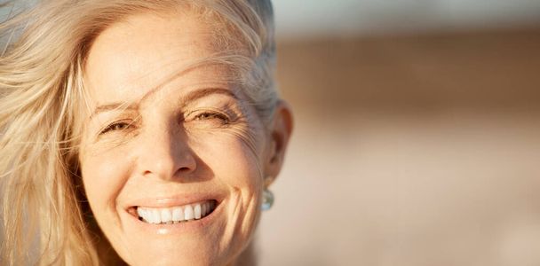 Face of beautiful mature woman on the beach. Smiling senior woman enjoying a beach holiday. Closeup on face of happy senior woman by the ocean. Portrait of a happy woman enjoying a vacation by the sea - 写真・画像