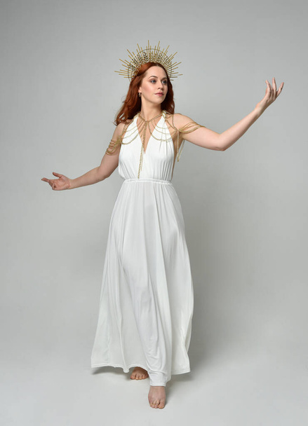 portrait of beautiful red head woman wearing long flowing fantasy toga gown with golden halo crown jewellery,  posed on isolated on a white studio background. - Φωτογραφία, εικόνα