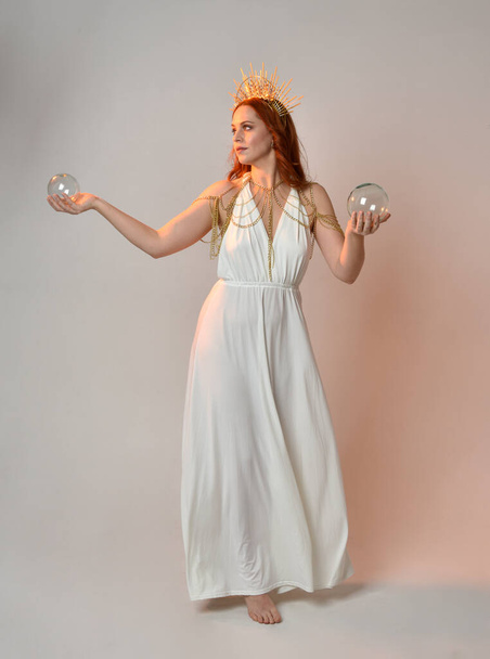 portrait of beautiful red head woman wearing long flowing fantasy toga gown with golden halo crown jewellery,  posed on isolated on a white studio background. - Valokuva, kuva