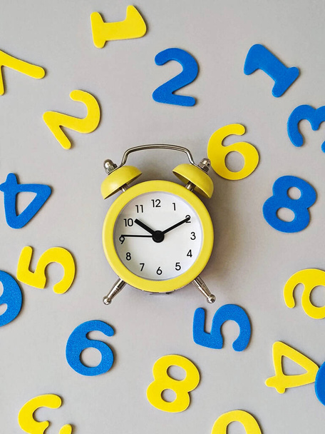 Illuminated yellow alarm clock on ultimate gray background with blue and yellow numbers flat lay. Back to school concept - Photo, Image