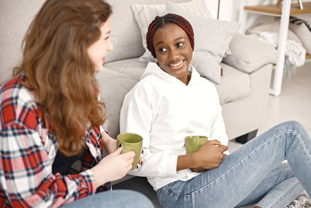 Two young woman sitting on a floor near sofa and talking. Black girl wearing white hoodie, caucasian girl wearing plaid shirt. Girls holding a green cups with tea. - Fotografie, Obrázek