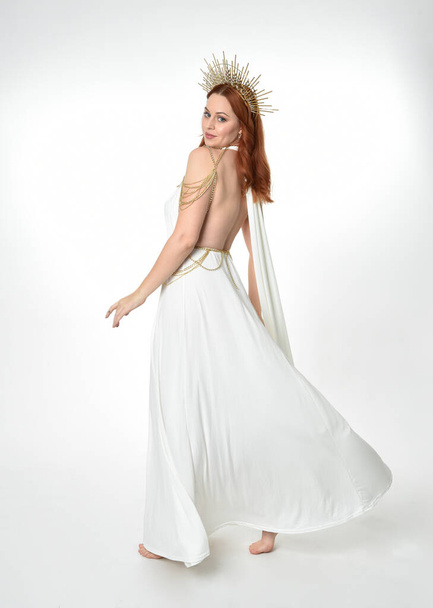 portrait of beautiful red head woman wearing long flowing fantasy toga gown with angelic  golden halo crown on studio background - Photo, image