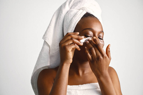 Black young woman standing isolated on white background with patches on her face. Girl has her hair wrapped in a towel. Black woman enjoy after body treatment. - Photo, image