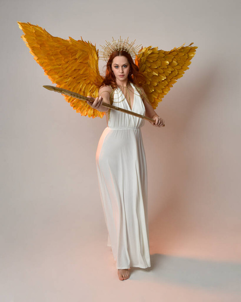 portrait of beautiful red head woman wearing long flowing fantasy toga gown with angelic  golden halo crown on studio background - Photo, Image