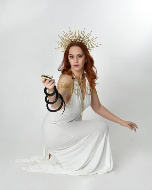 ritratto di bella donna testa rossa che indossa lungo fluente fantasia toga abito con angelico aureola corona d'oro su sfondo studio - Foto, immagini