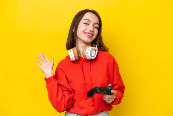 Young Ukrainian woman playing with a video game controller isolated on yellow background saluting with hand with happy expression - Фото, зображення