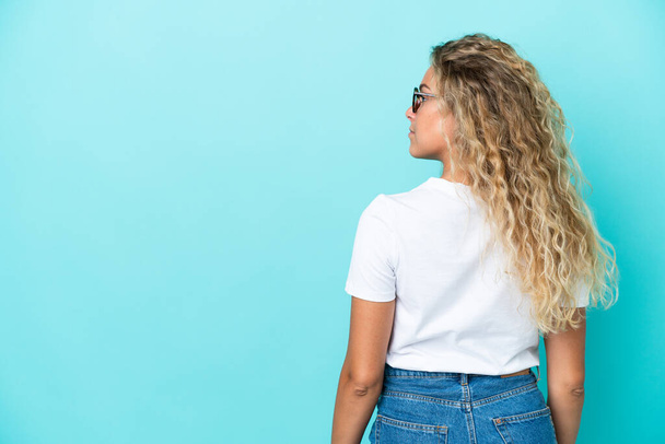 Girl with curly hair isolated on blue background in back position and looking side - Φωτογραφία, εικόνα