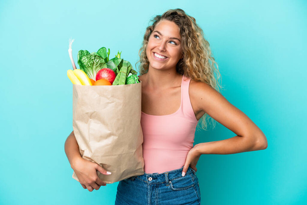 Dziewczyna z kręconymi włosami trzymając torbę na zakupy spożywcze izolowane na zielonym tle pozowanie z ramionami na biodro i uśmiech - Zdjęcie, obraz