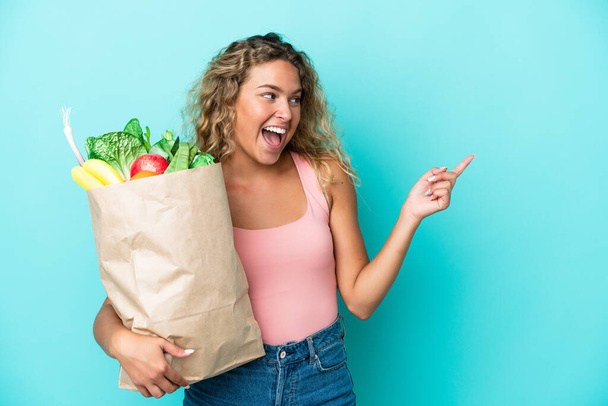 Girl with curly hair holding a grocery shopping bag isolated on green background pointing finger to the side and presenting a product - Photo, Image